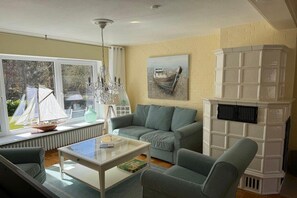 Ein Wohnzimmer mit einer Couch, einem Sessel und einem Glastisch. Ein Kamin mit weißem Sims und ein Gemälde an der Wand sind ebenfalls zu sehen.