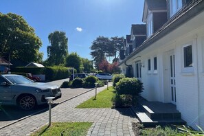A residential street with parked cars, a white building with a porch, and a clear sky. - Holiday apartment with 1 bedroom (Hohwacht)