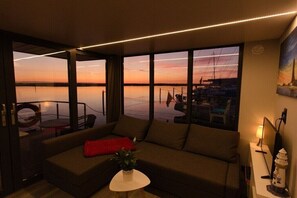 Sala de estar moderna com uma grande janela para um porto deportivo ao pôr do sol.