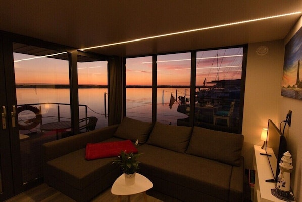 A modern living room with a large window overlooking a marina at sunset. - Houseboat in Wendtort with swimming pool (Wendtorf)