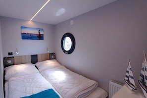A bedroom with a bed, a round window, a painting of a lighthouse, and a small sailboat model.