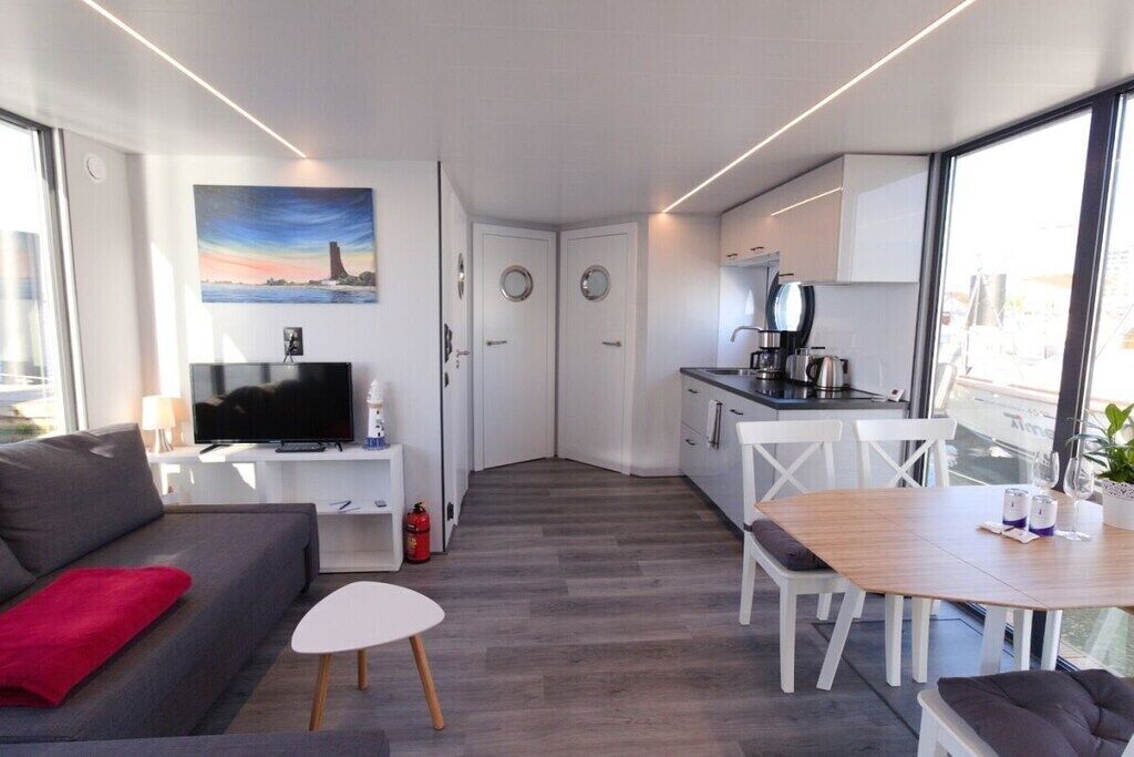 Ein gemütlicher Wohnraum mit einer Couch, einem kleinen Tisch, einem Fernseher, einer Küchenzeile und einem großen Fenster.