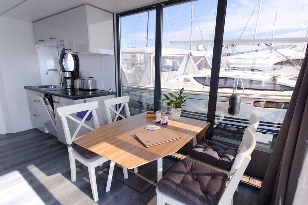 A compact kitchen with modern appliances, a dining area with a wooden table and white chairs, and a view of boats through large windows.