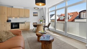 Eine moderne Küche mit Holzausstattung, ein Essbereich mit rundem Tisch und Stühlen sowie große Fenster mit Blick auf Häuser mit roten Dächern.