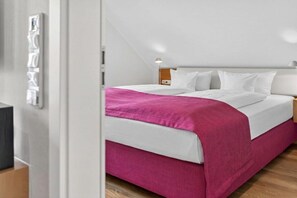 Une chambre à coucher avec un lit, du linge de lit blanc, une touche de rose, une table de chevet et une lampe.