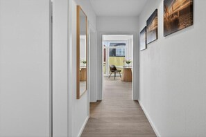 Un couloir doté d’un plancher en bois, de murs blancs et d’une porte donnant sur une pièce où se trouvent une chaise et une table.