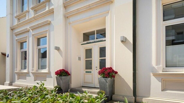 Ein Gebäude mit weißer Fassade, einer Tür mit der Aufschrift 'Maison Christine' und zwei großen Blumenkästen.