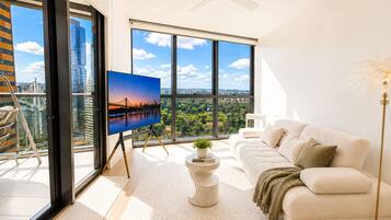 A modern living room with a large window, a white sofa, a coffee table, and a television displaying a cityscape.