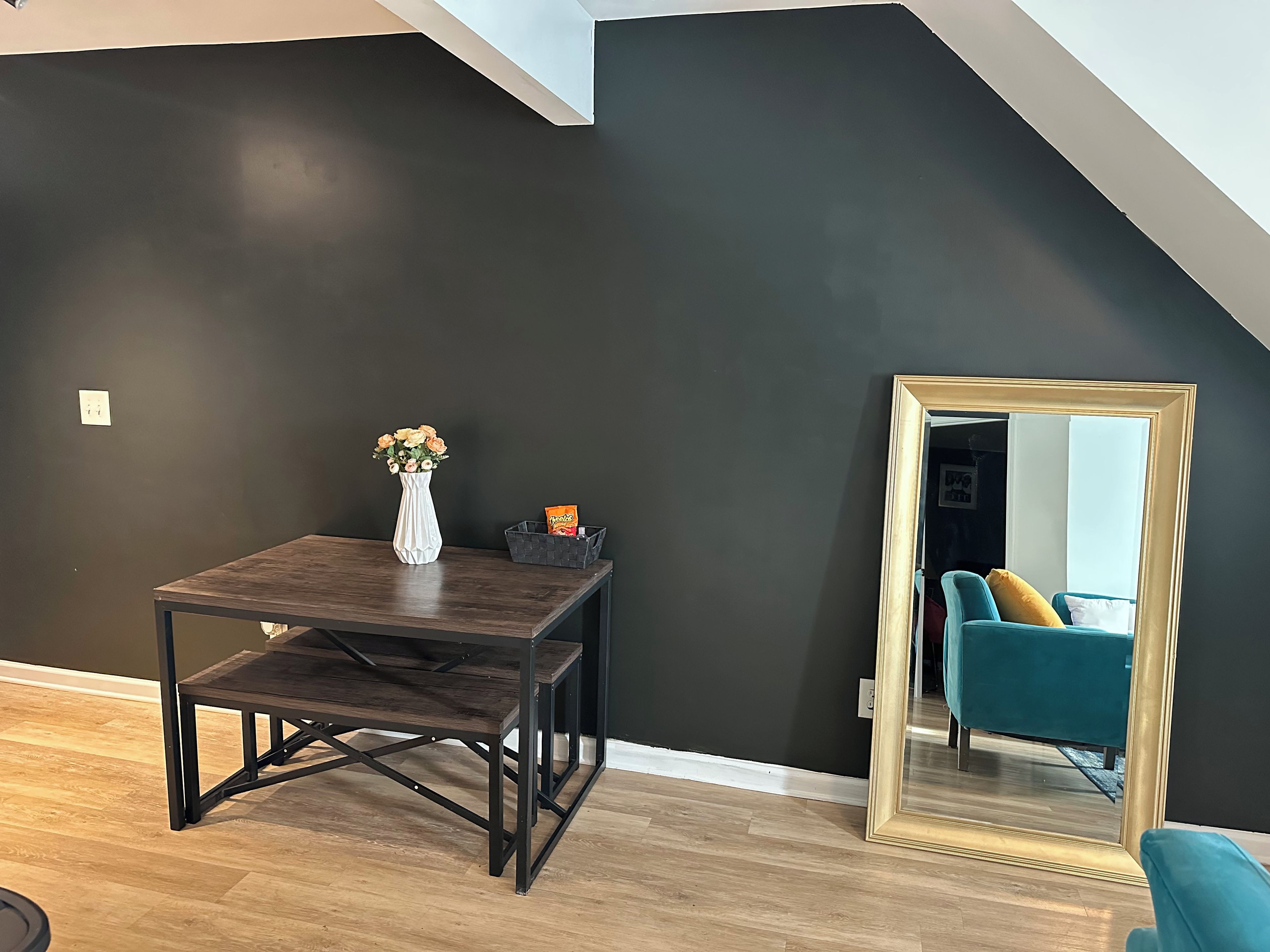 A room with a dark wall, a wooden table with a vase and snacks, a mirror, and a teal chair.