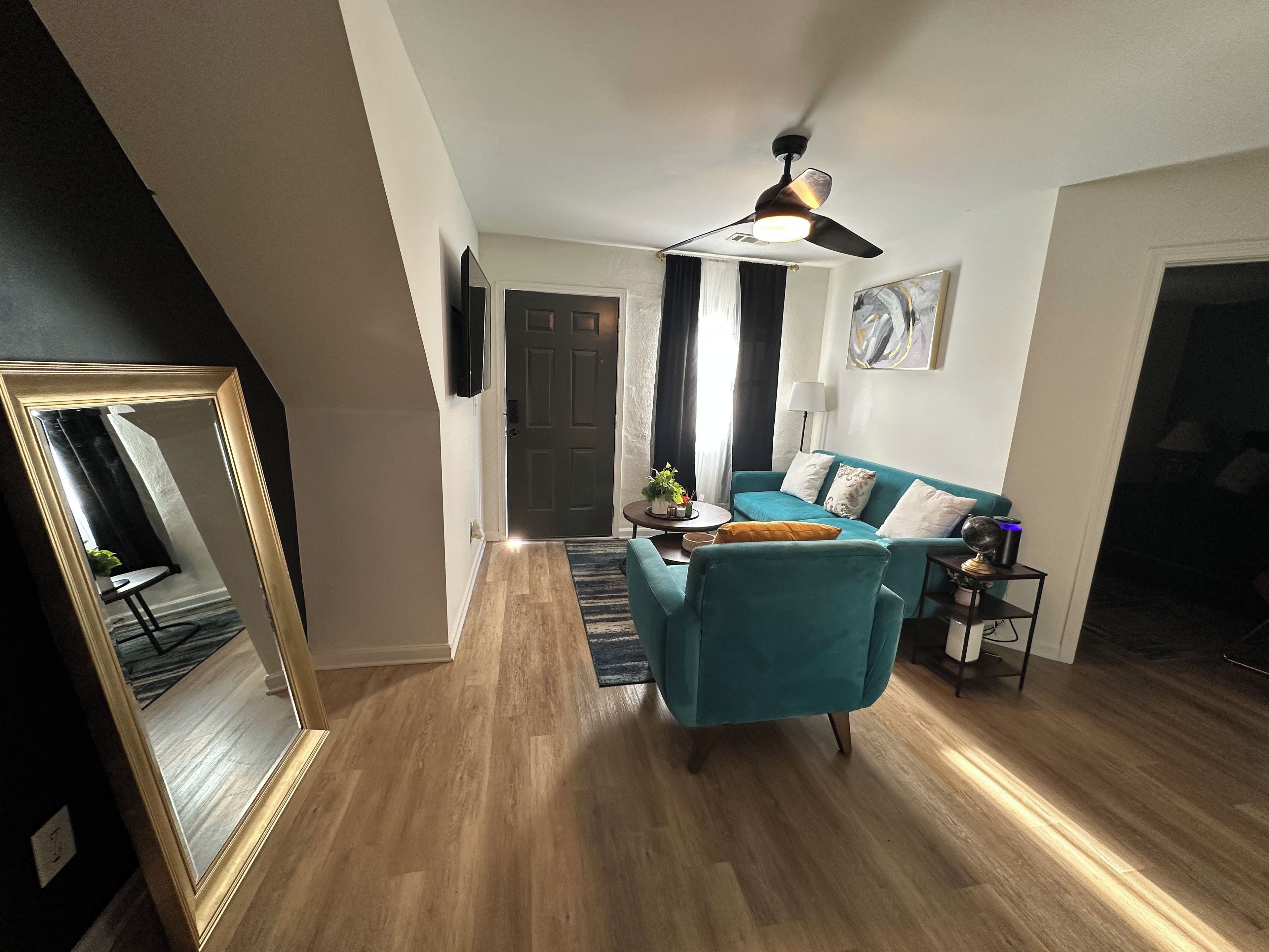 A living room with a teal sofa, a ceiling fan, a TV mounted on the wall, and a wooden floor.