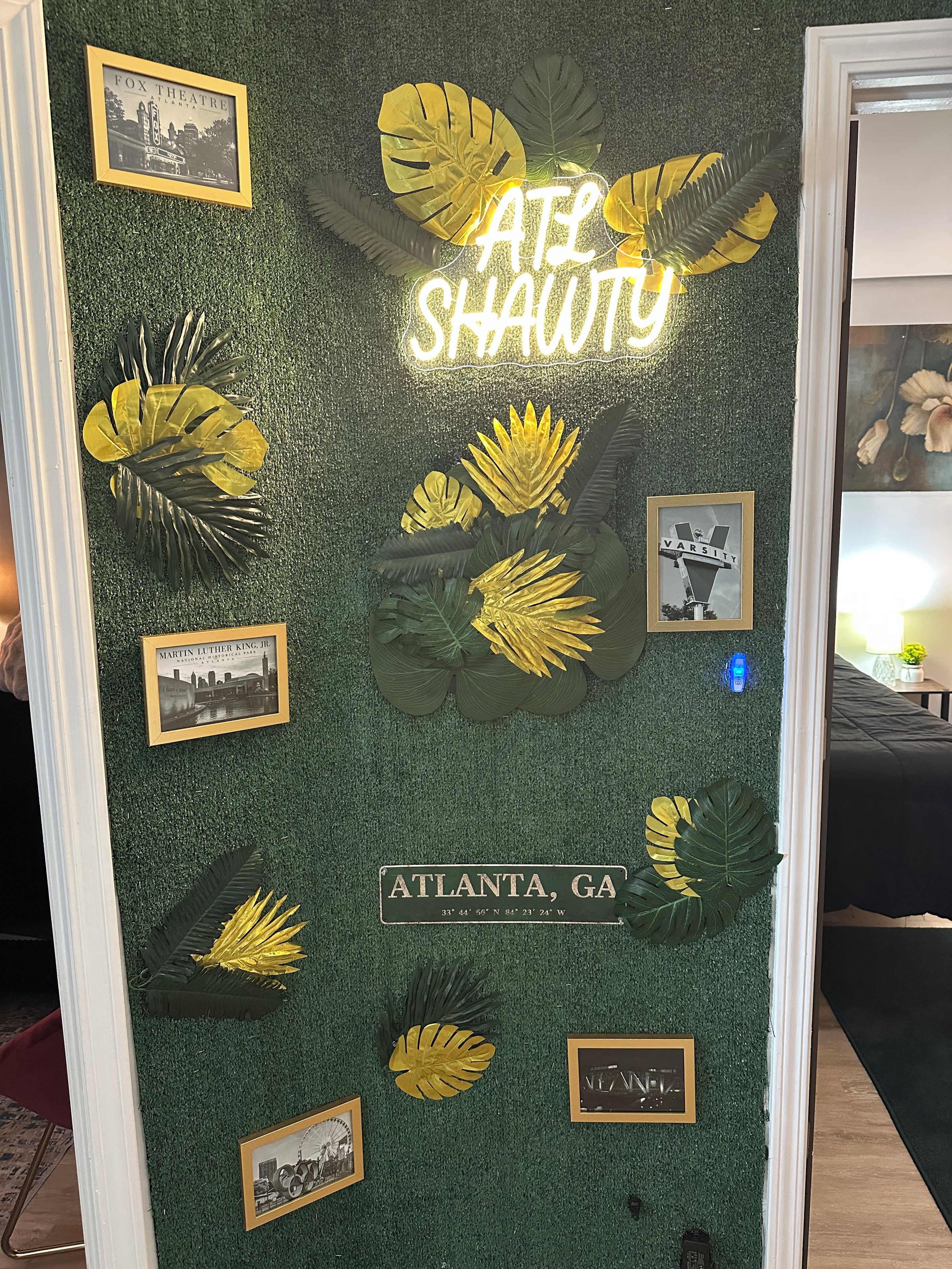 A wall with framed pictures, a neon sign, and decorative leaves.