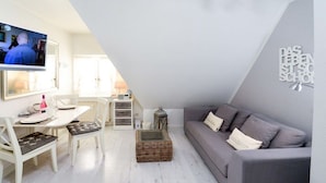 A modern living room with a grey sofa, white dining table, and a television mounted on the wall. - Apartment in Rantum with swimming pool (Rantum)