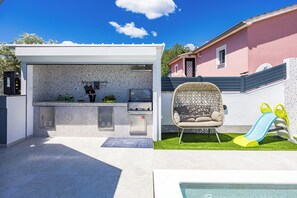 An outdoor kitchen area with a barbecue grill, a seating area with a wicker chair, and a children's slide. - Villa Spa Otium by Istra Vacation (loborika)