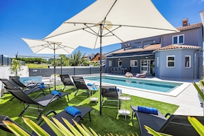 Een zwembadgedeelte met ligbedden, een grote parasol en een modern huis op de achtergrond.