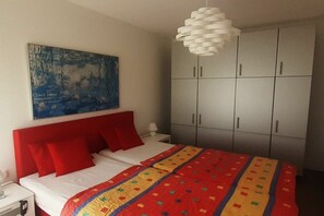 Une chambre à coucher avec un lit, des oreillers rouges, une couverture colorée, un luminaire blanc au plafond, une armoire grise et un tableau accroché au mur.