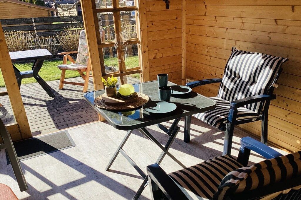 A wooden cabin with a glass table and striped chairs, a small garden visible through the window, and a wooden deck.
