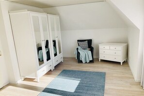 A room with a white wardrobe, a small chair with a cushion, a white dresser, and a blue and white rug on a wooden floor. - Holiday apartment with 1 bedroom (Maasholm)