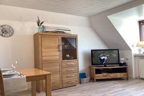 A room with a wooden cabinet, a television, a clock, and a dining table set with plates and a glass. - Holiday apartment with 1 bedroom (Maasholm)