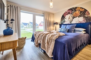 A bedroom with a large bed, a wooden table, a vase with flowers, and a view of houses through the window. - Holiday apartment in Büsum with swimming pool (Büsum)
