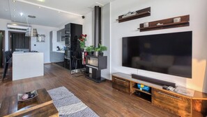 A modern living room with a flat-screen TV, wooden entertainment unit, and a kitchen area in the background.