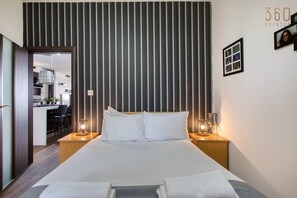 A hotel room with a large bed, two bedside tables with lamps, a striped headboard, and a kitchenette visible through an open door.
