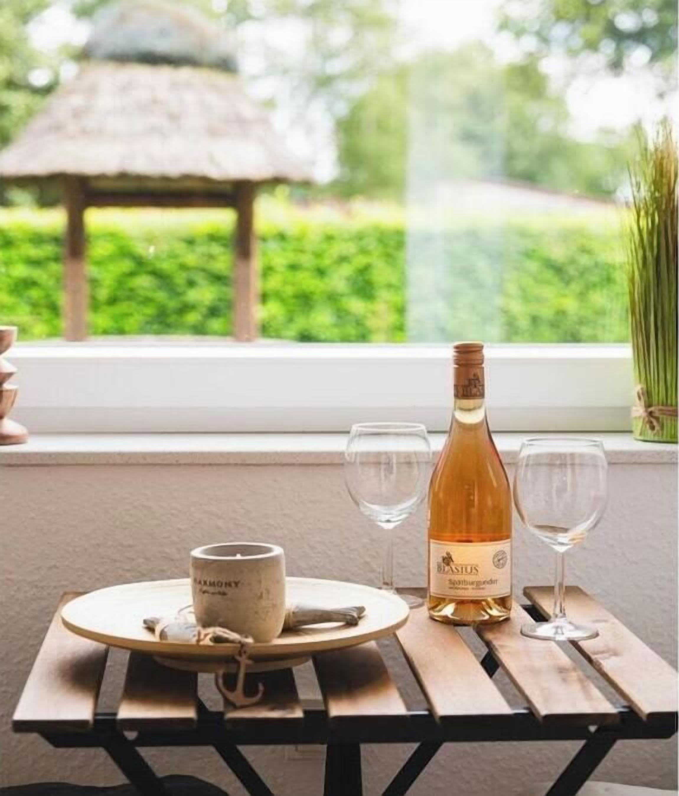 A table with a bottle of wine, two glasses, and a mug on a wooden table near a window with a view of a garden.