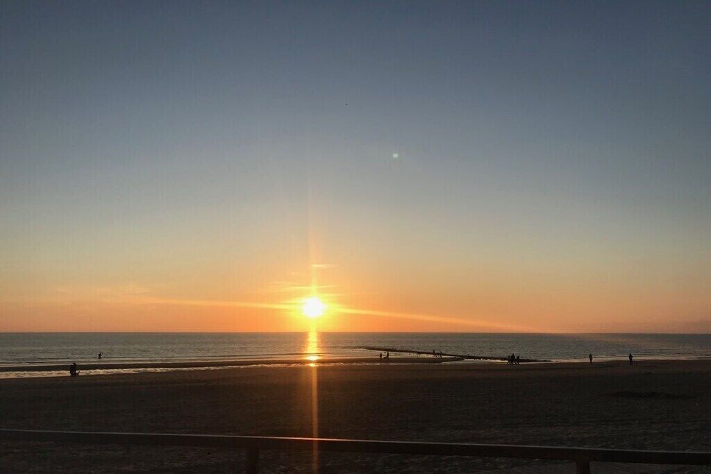 A beach scene with the sun setting over the horizon, a clear sky, and a calm sea.