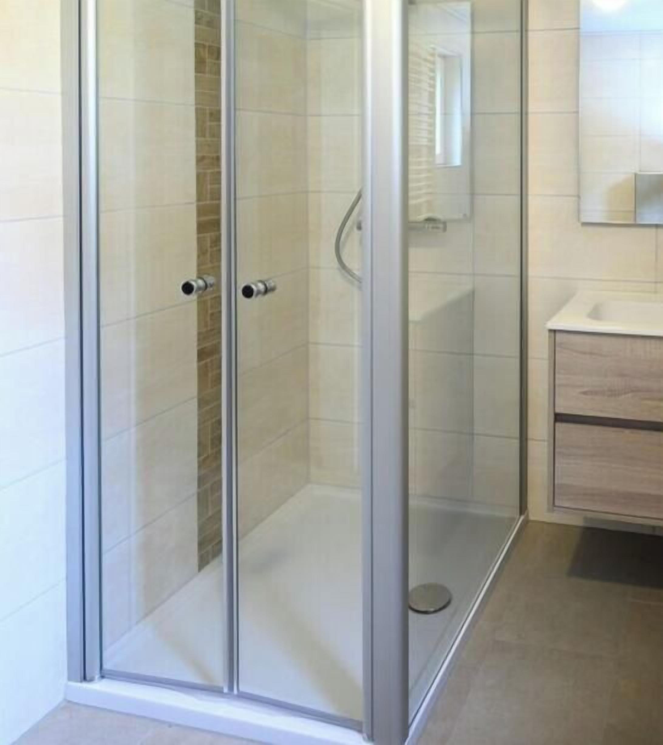 Une douche fermée par des parois de verre, avec un mur recouvert de carreaux blancs et un meuble-lavabo en bois doté d’un miroir.