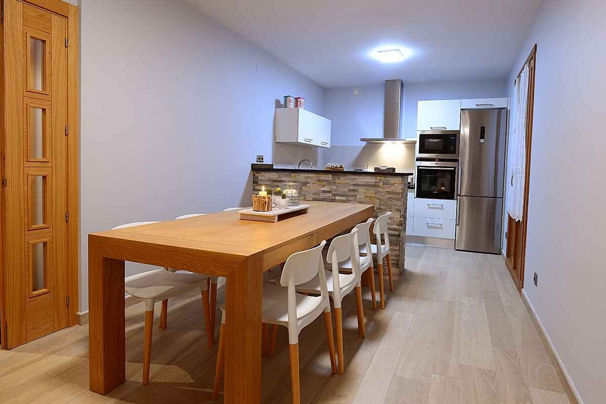 Cocina compacta con mesa de comedor de madera, sillas blancas, horno empotrado y refrigerador de acero inoxidable.