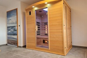 Eine Sauna mit Holzfassade und Glastür, in einem Raum mit gefliestem Boden und einem Wandbild mit Strandmotiv.