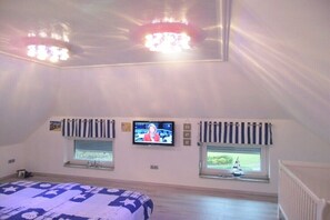 Un dormitorio con cama, dos ventanas con cortinas, televisión y lámpara de techo.