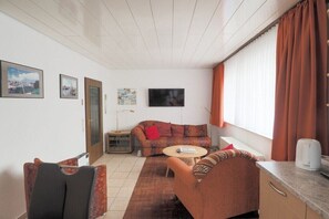 Een woonkamer met een bank, fauteuil en een klein tafeltje. Aan de muur hangt een televisie en er is een raam met gordijnen.