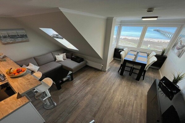 Un espai habitable compacte amb zona de menjador, sofà, cuina petita i vistes al mar a través de grans finestres.