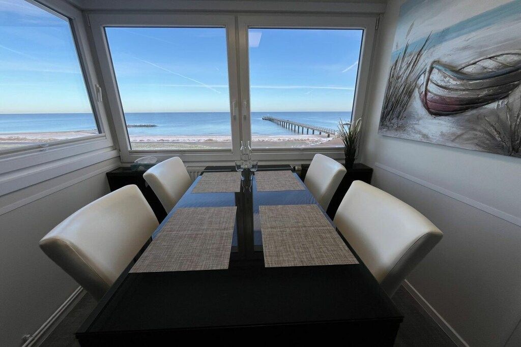A dining area with a view of the beach and pier, featuring a table with four chairs and a painting on the wall.