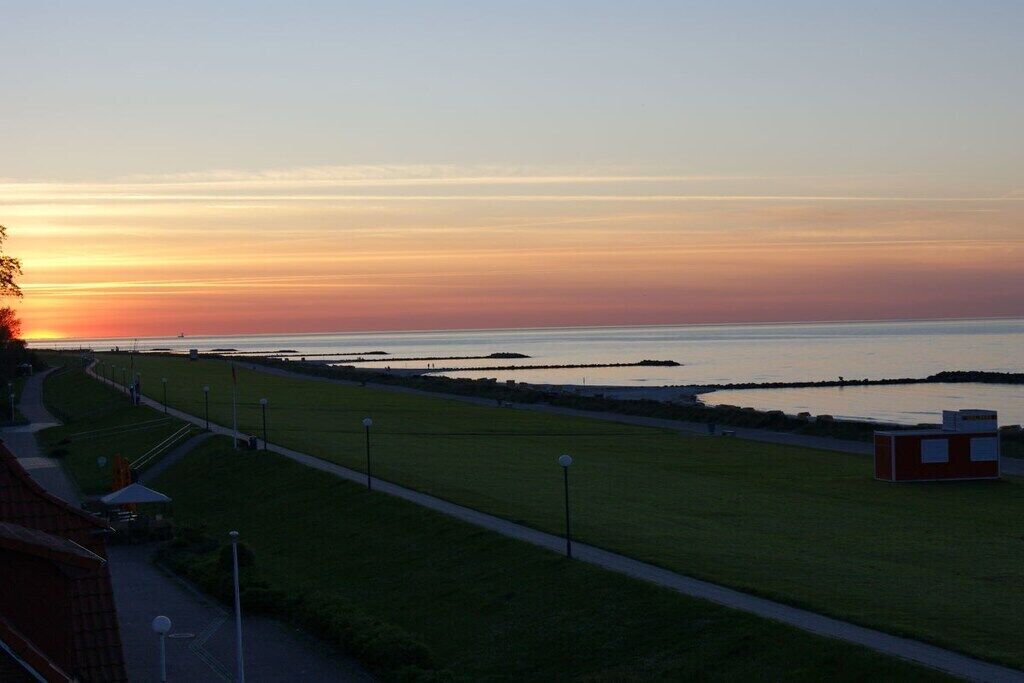 Sonnenuntergang über einer Küstenlandschaft mit einem grasbewachsen Pfad, einem kleinen Gebäude und einem Gewässer.