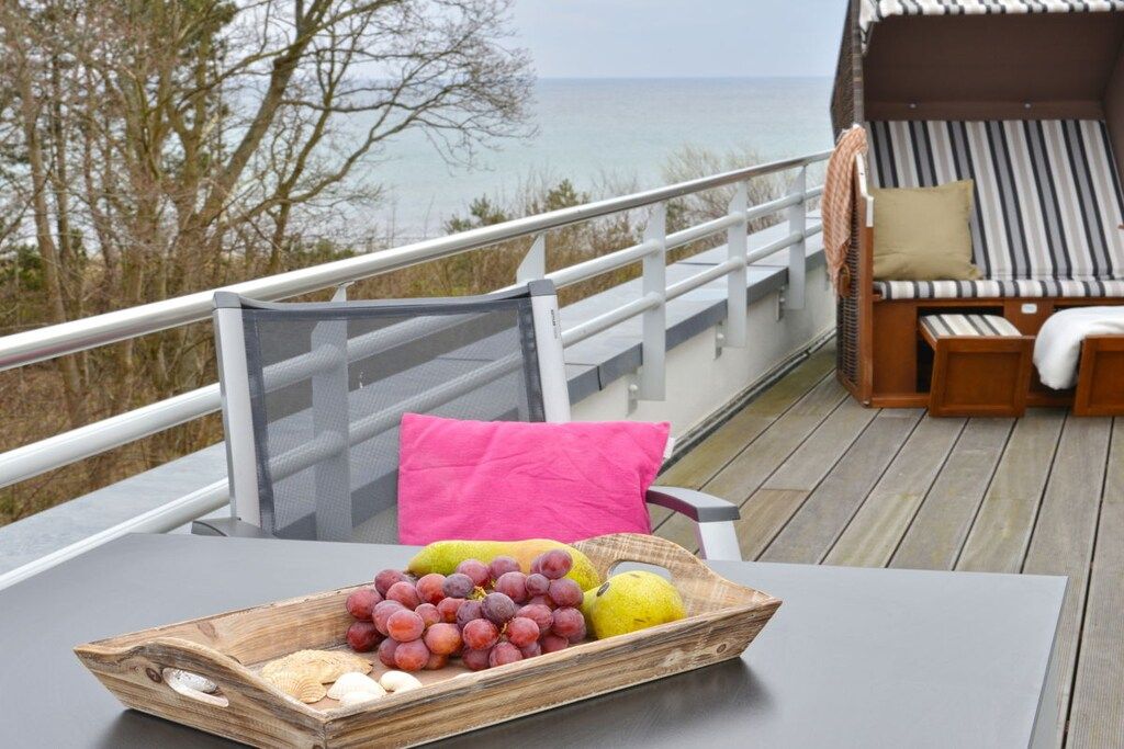 Una terraza con una mesa que sostiene una bandeja de frutas, un área de estar con cojines y vistas al mar, y una barandilla.