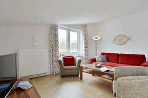 Ein Wohnzimmer mit einem roten Sofa, einem Holztisch, einem Fernseher und einem Fenster mit blumigem Vorhang.