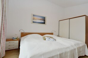 A bedroom with a bed, bedside table, lamp, and a view of the sea through a window. - Holiday apartment with 1 bedroom (Breege\/Juliusruh OT Juliusruh)