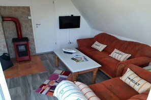Ein Wohnzimmer mit einer Couch, einem an der Wand montierten Fernseher, einem kleinen Tisch mit Zeitschriften und einem Kamin mit rotem Schornstein.
