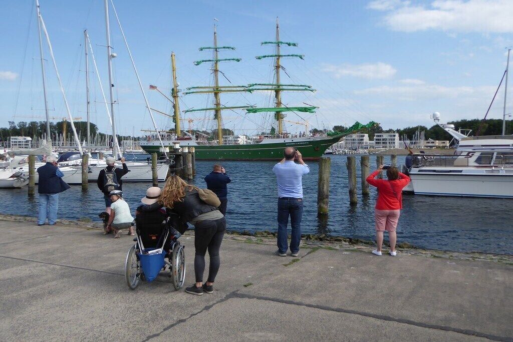 A marina with a large sailing ship docked, surrounded by people, some taking photos, and a wheelchair user present.