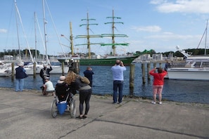 Ein Yachthafen mit einem großen Segelschiff, umgeben von Menschen, von denen einige Fotos machen, und einer Person im Rollstuhl.