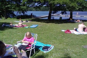 Ein Park mit Menschen, die auf der Wiese liegen und sich entspannen, ein See im Hintergrund und ein Picknicktisch.