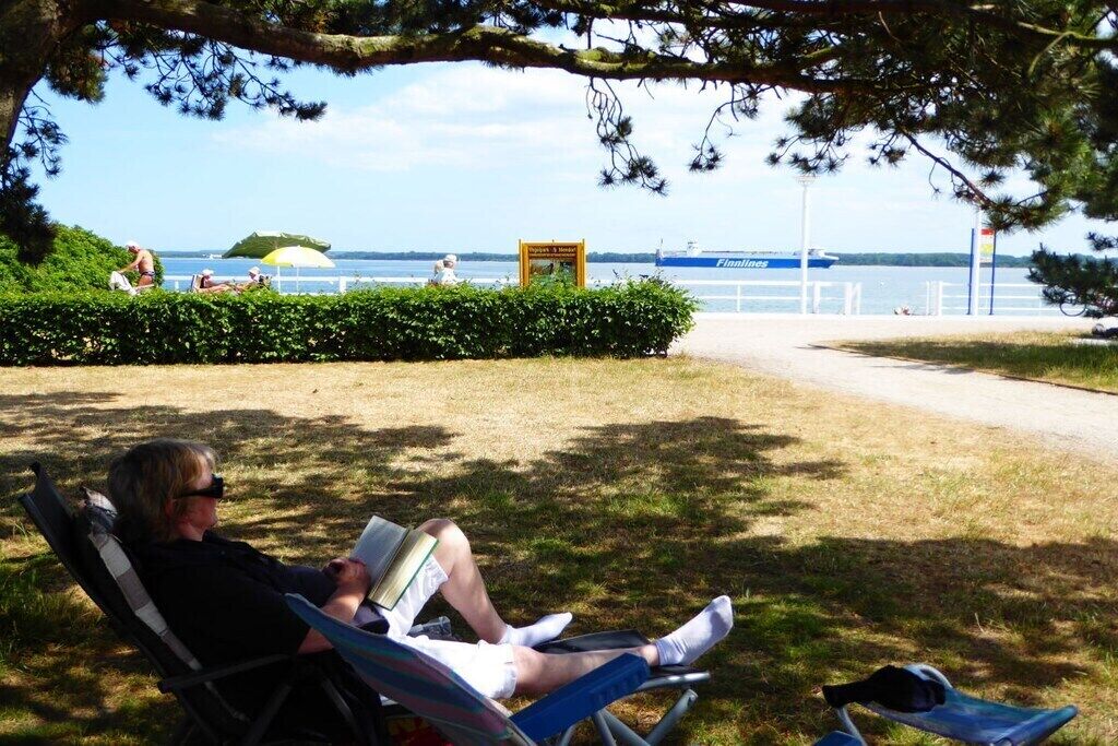 A park with a view of a body of water and a boat. There are people sitting on chairs and a bench.