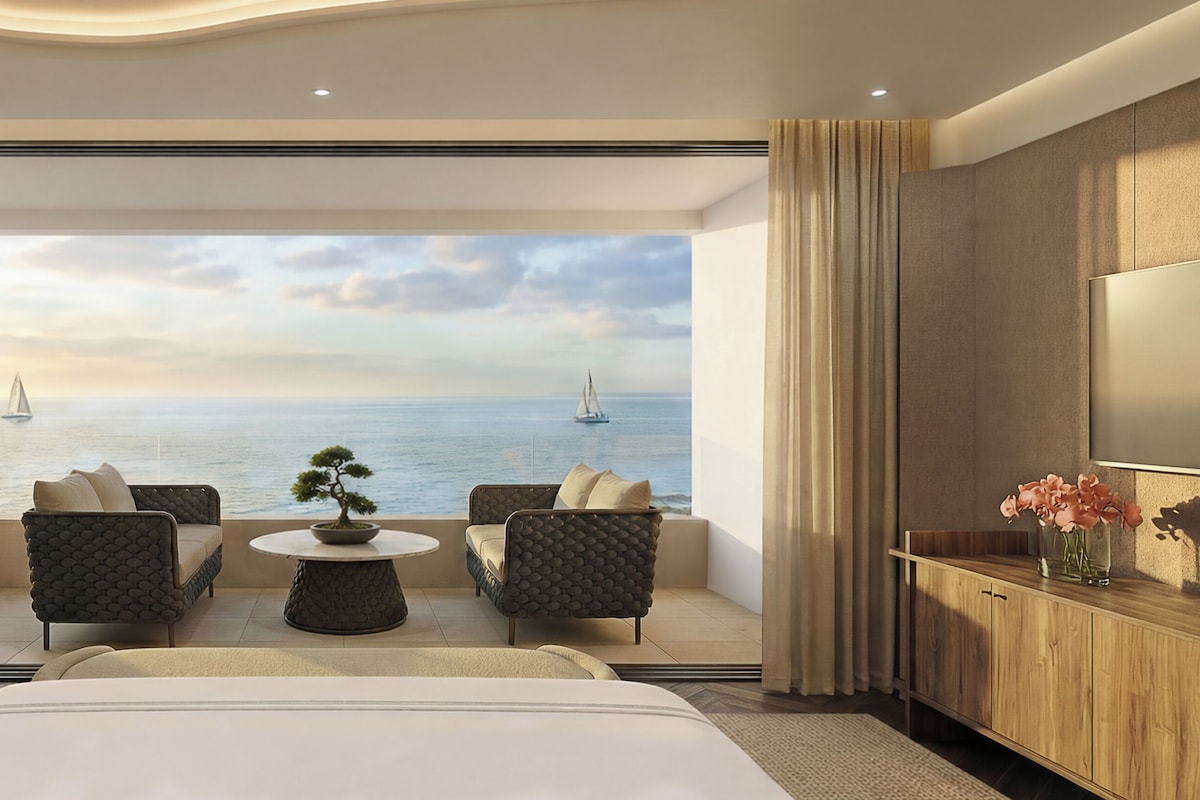 Une chambre d’hôtel avec un grand lit, deux fauteuils, une petite table, une plante en pot et une vue sur la mer, où l’on peut apercevoir des voiliers.