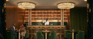 Un espace bar avec un barman, des étagères en bois remplies de bouteilles et des chaises recouvertes de tissu vert.