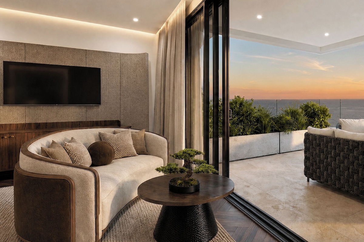 Un salon moderne avec un canapé, des fauteuils, une table basse et un téléviseur fixé au mur. Une grande fenêtre offre une vue sur l’océan et sur le coucher de soleil.