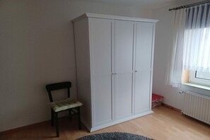 Ein Zimmer mit weißem Kleiderschrank, einem kleinen Stuhl, einem Heizkörper und einem Fenster mit Vorhängen.