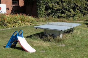 Ein Pingpongtisch in einer grünen Wiese mit einer Rutsche und einem Backsteinhaus im Hintergrund.
