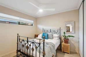 A bedroom with a bed, a ceiling fan, a window, a bedside table with a plant, and a mirror on the wall. - Terra de Sol | Terracotta Courtyard, Swan Valley (Bassendean)