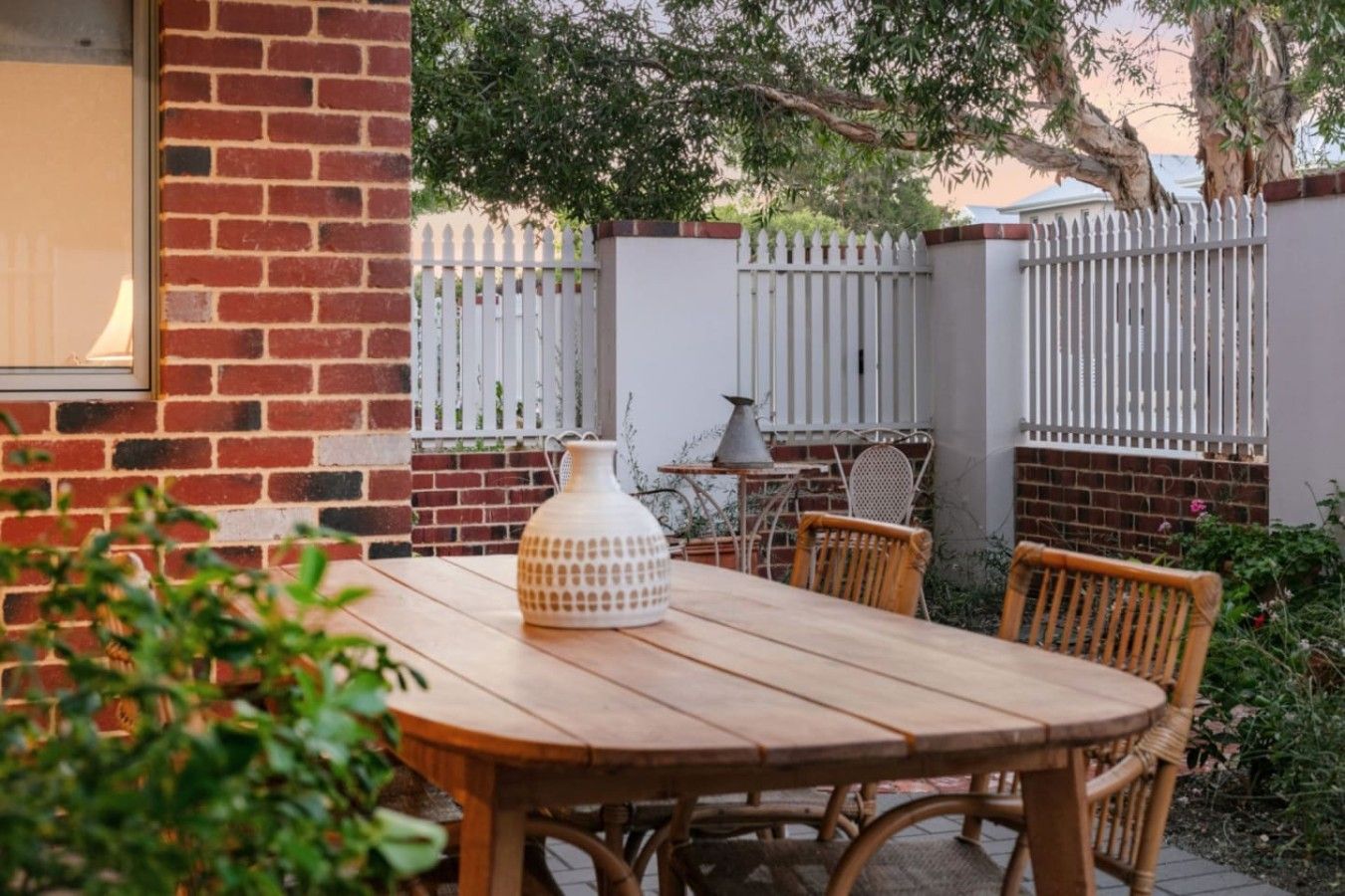 A wooden table with chairs, a white vase, and a metal lamp outside a brick building with a white picket fence.